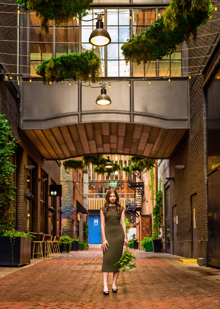 Woman in an olive-green dress stands in a brick alley lined with hanging plants and warm string lights under a glass canopy.