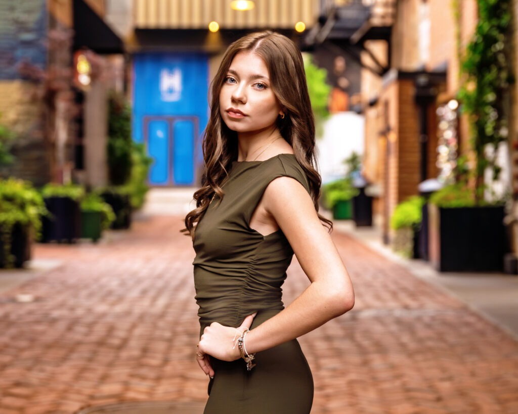 Portrait of a woman in an olive-green dress posing on a cobblestone street with blurred storefronts behind her