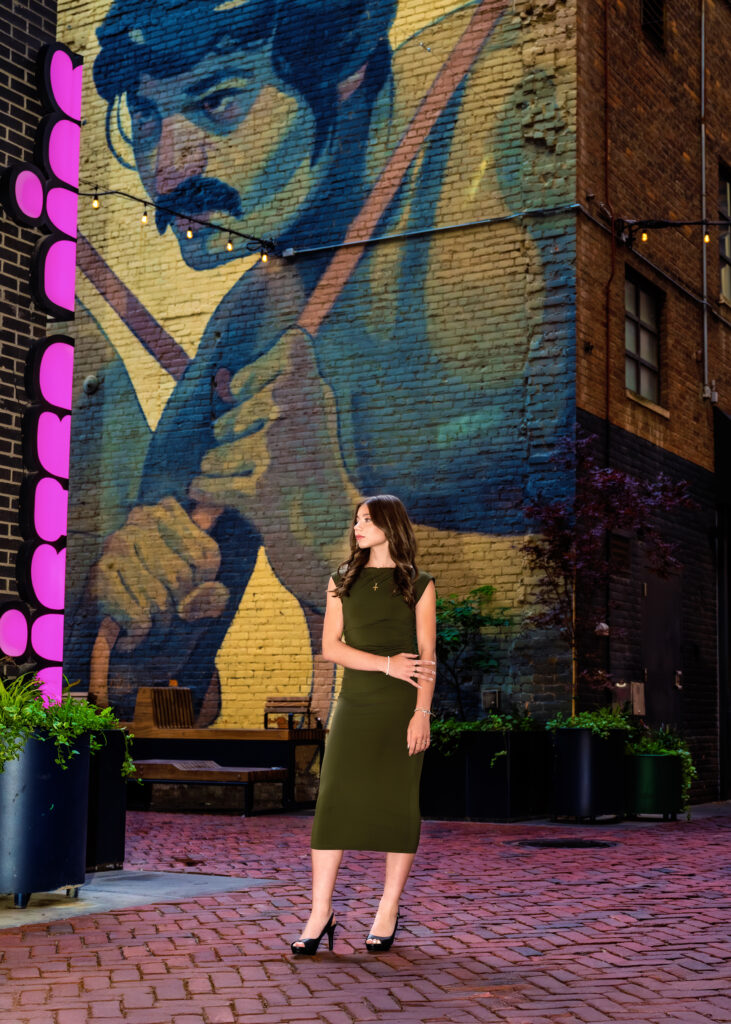 Woman in an olive-green dress posing in front of a large brick-wall mural with string lights overhead and pink neon signage to the left.