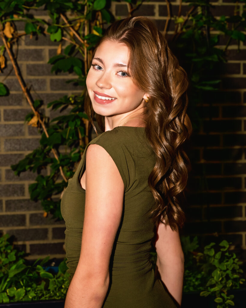 Smiling young woman with long wavy brown hair in an olive-green dress, standing against a brick wall with green plants behind her.