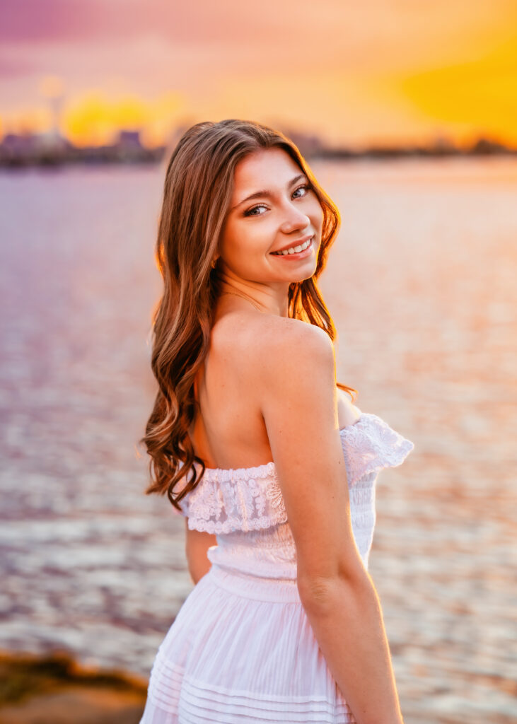 Smiling woman in a white off-shoulder dress stands by a sunset-lit sea.