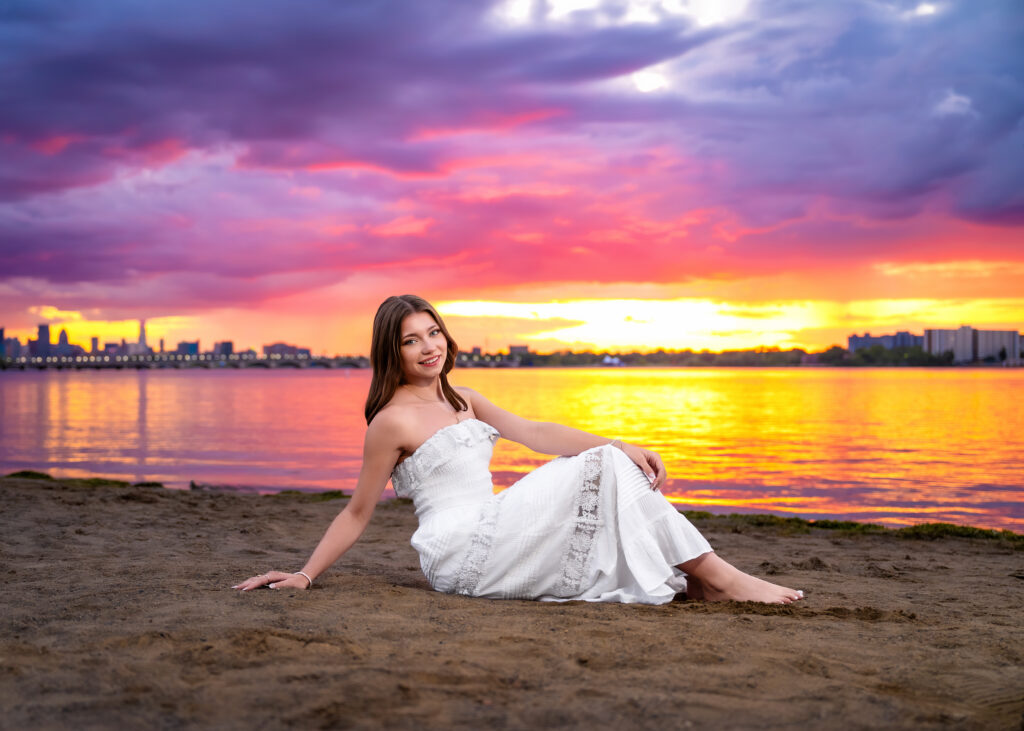 Woman in a white dress sits on a sandy beach at sunset, with a colorful purple-pink-orange sky over the water and distant city skyline on the horizon.