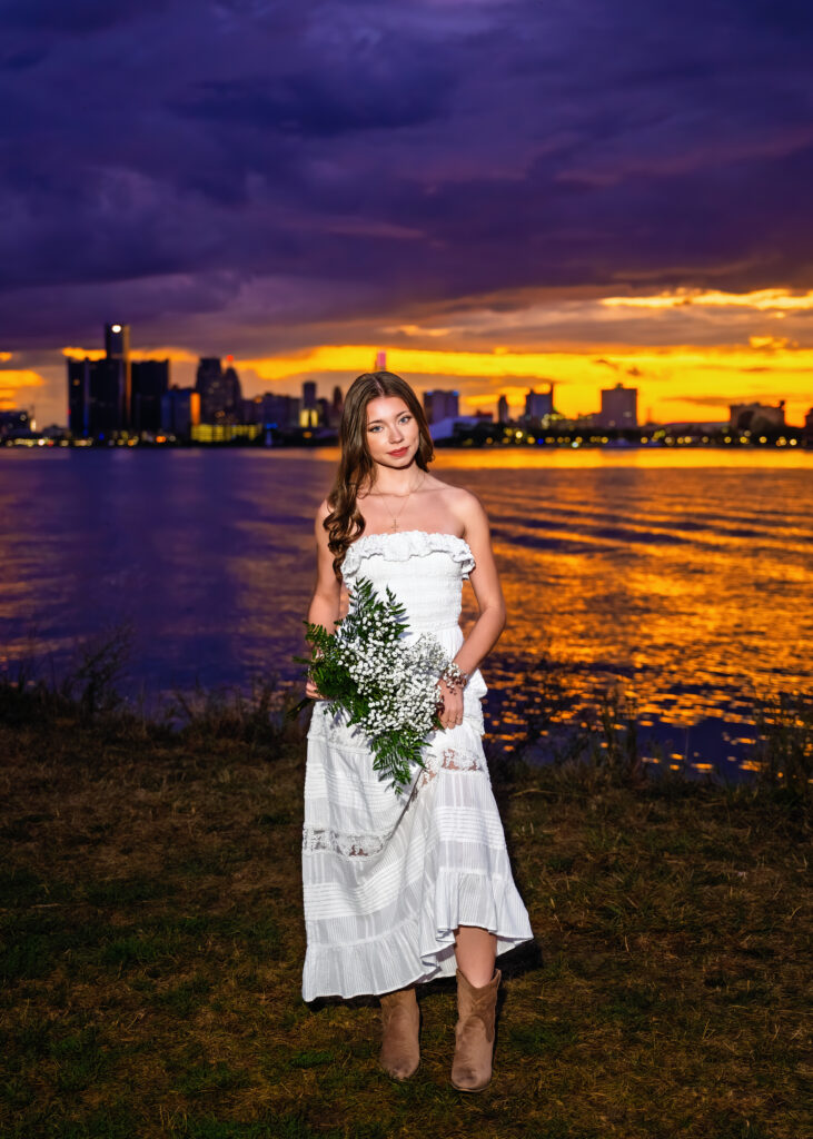Woman in a white dress holding a bouquet of white flowers by a lake with a city skyline at sunset behind her.