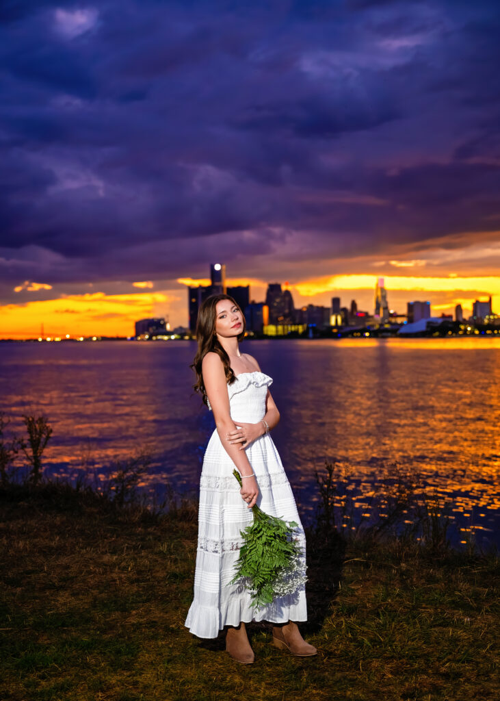 Woman in a white dress standing by the water, holding a fern, with a city skyline and orange sunset in the background.