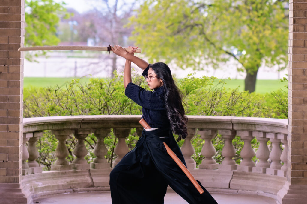 Woman in dark clothing practicing with a wooden sword overhead in a brick gazebo with green trees in the background.