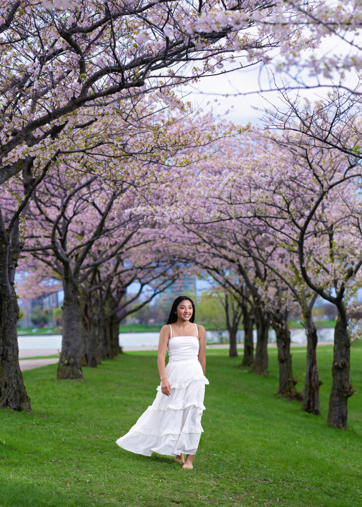 Woman in a white dress walking barefoot on grass under blooming cherry trees in a park."],