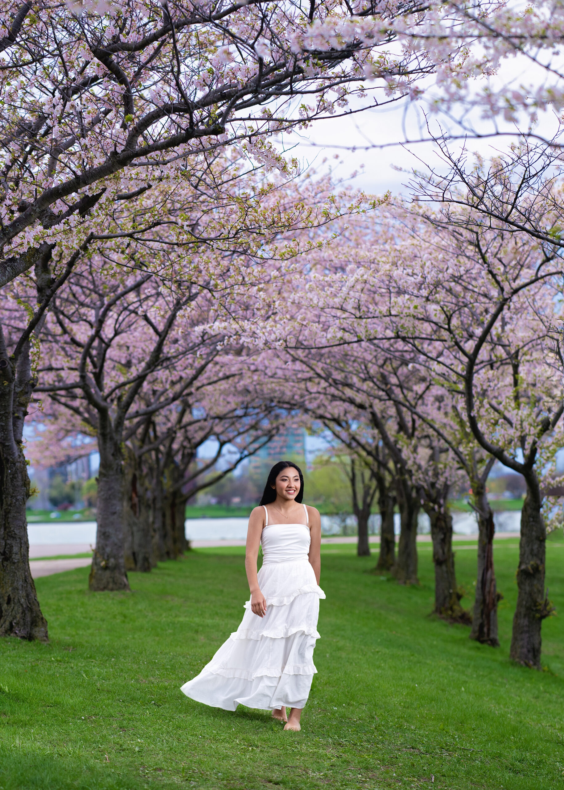 Woman in a white dress walking barefoot on grass under blooming cherry trees in a park."],