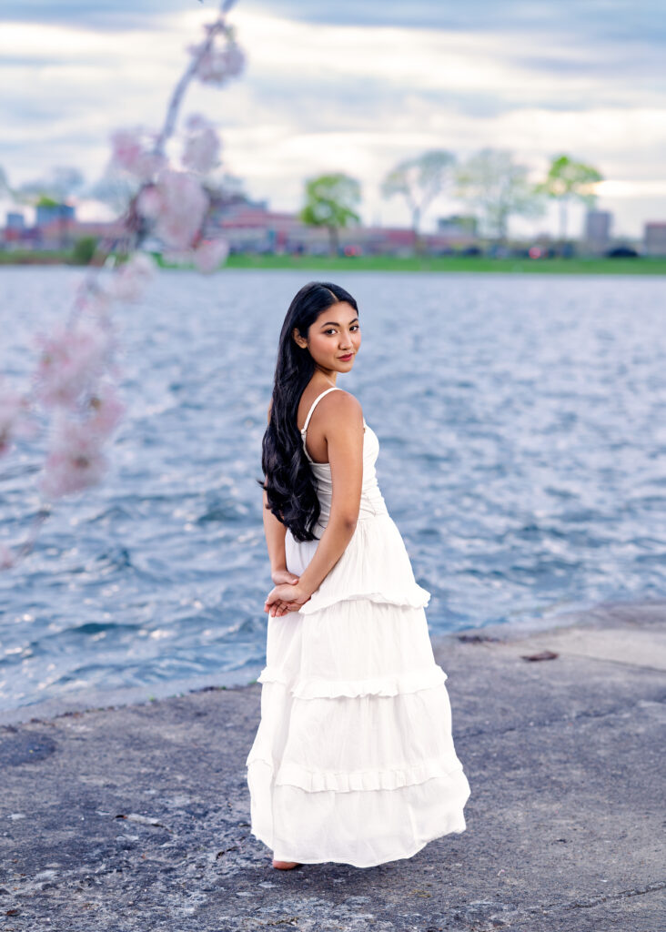 Woman in a white, tiered dress stands barefoot on a rocky lakefront, looking back over her shoulder toward the camera.