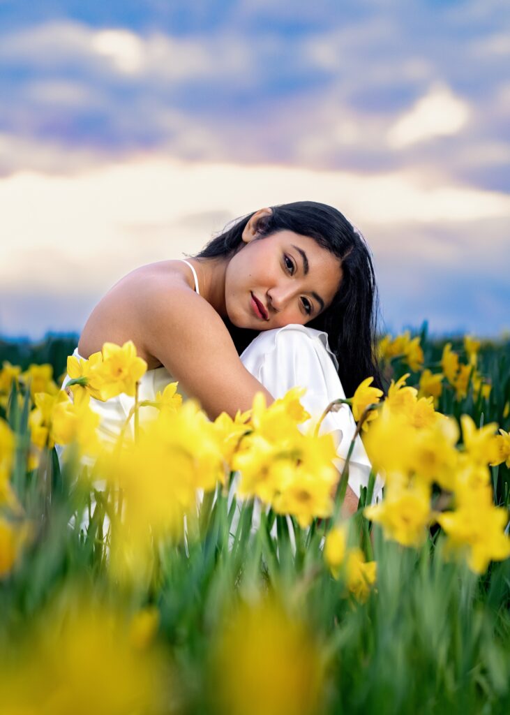 Smiling woman with dark hair in a white outfit leaning among bright yellow daffodils, blue sky with clouds in the background.|