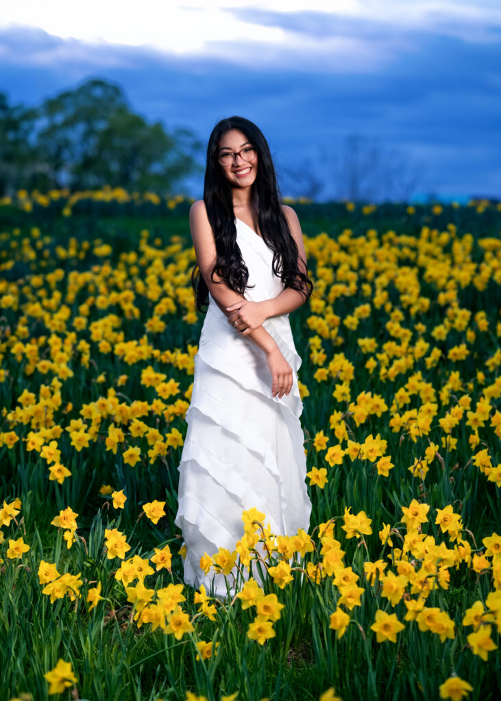 Smiling woman with long dark hair and glasses in a white dress stands in a field of yellow daffodils under a blue sky.